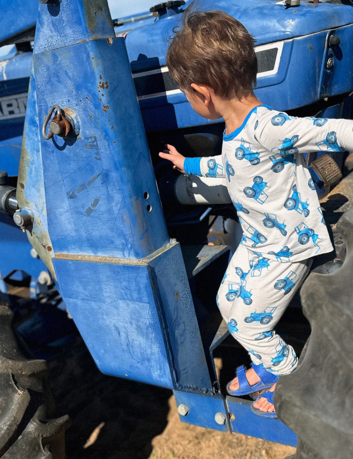 Child climbing on a blue tractor wearing a blue tractor design pajama set with a clear sky background