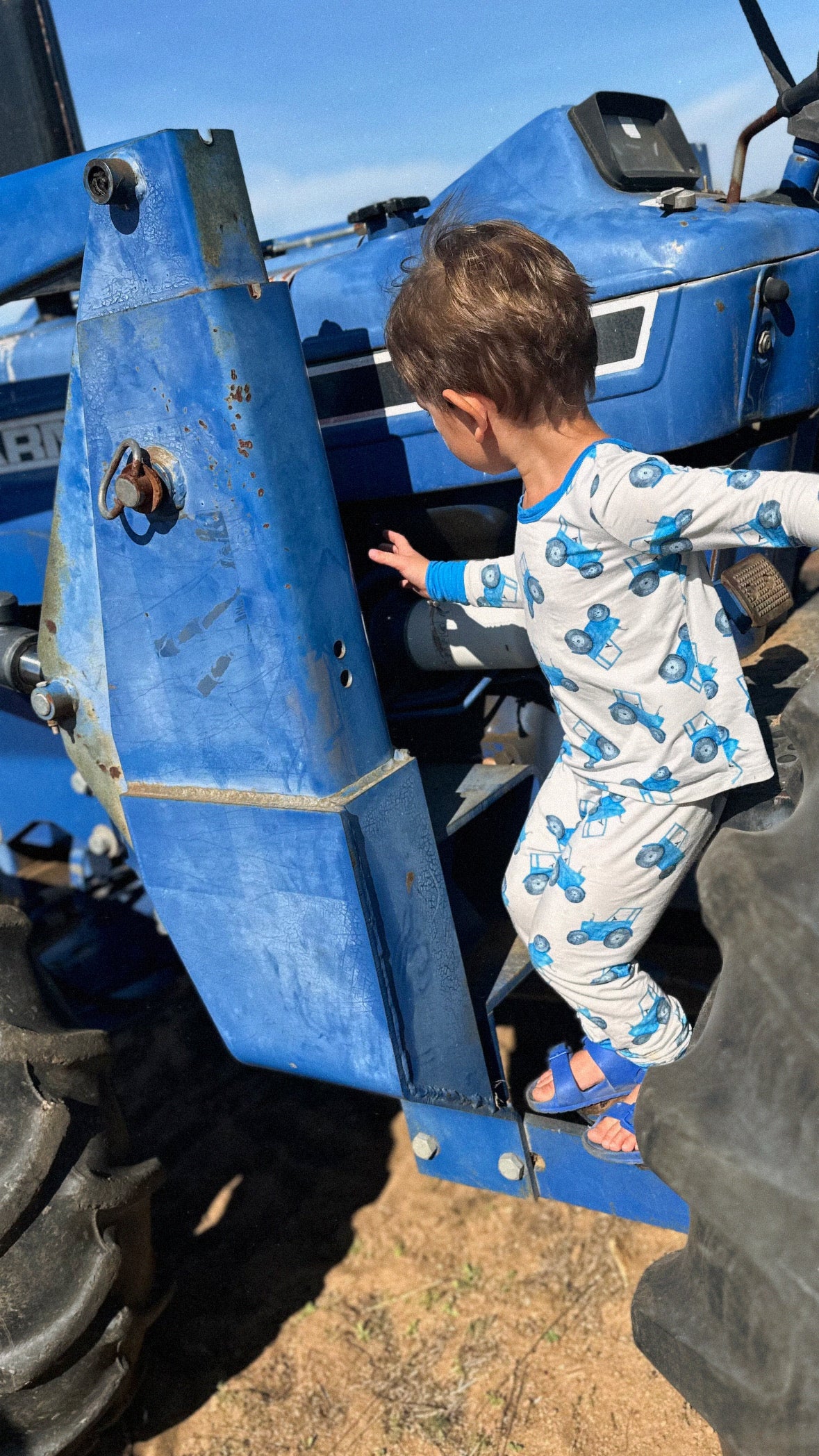 Child climbing on a blue tractor wearing a blue tractor design pajama set with a clear sky background