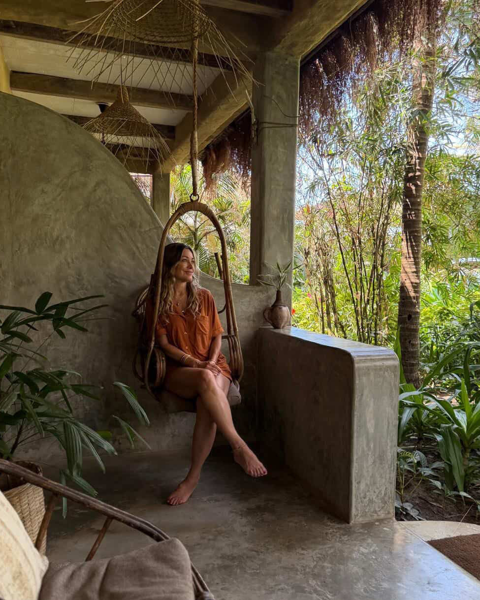 Woman sitting in a hanging chair on a tropical patio with lush greenery.