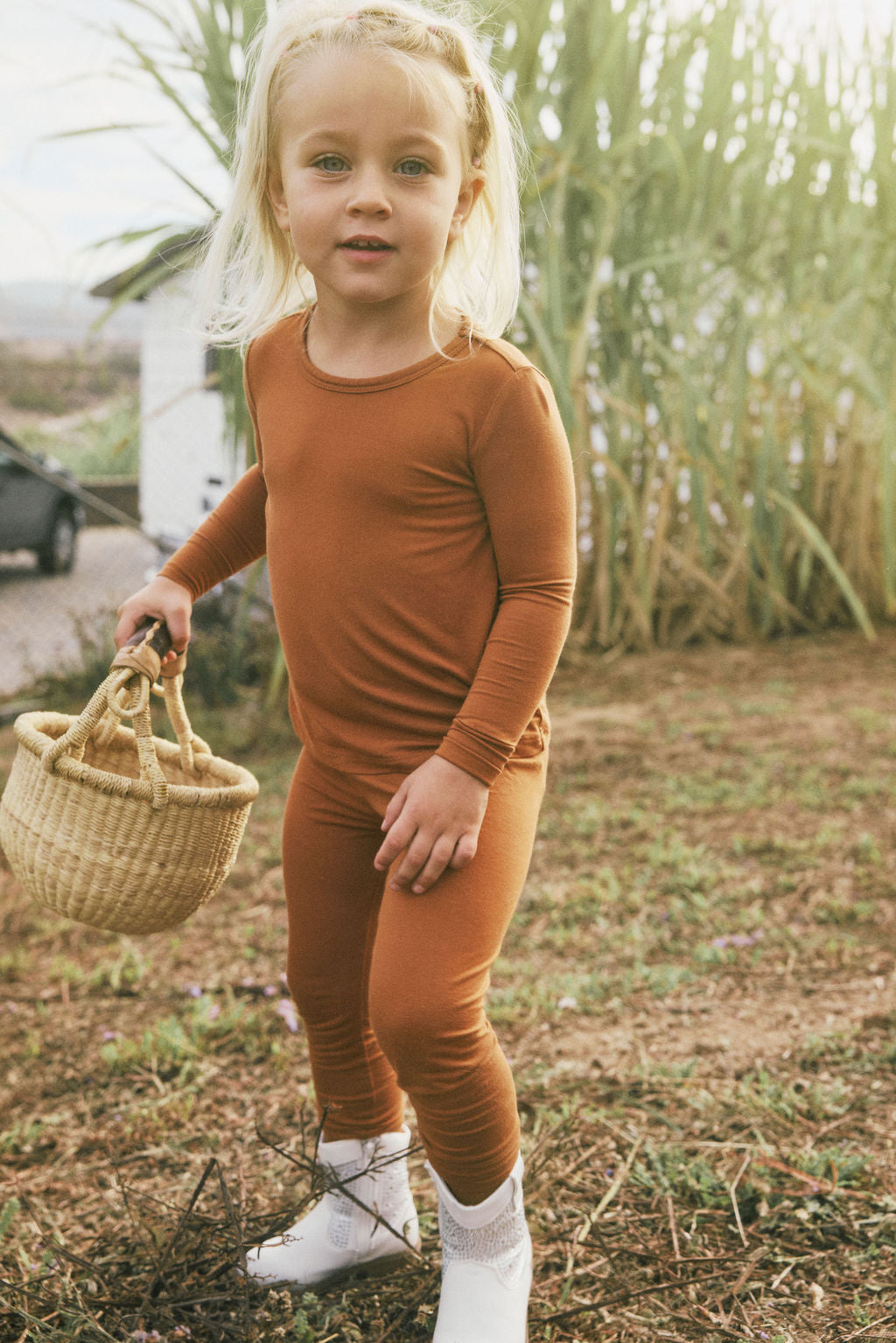 Child in a terracotta-colored outfit pajama set holding a woven basket outdoors.