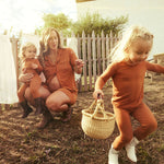 Woman and two children in matching terracotta pajamas outdoors with a wooden fence and clothesline in the background.