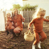 Woman and two children in matching terracotta pajamas outdoors with a wooden fence and clothesline in the background.