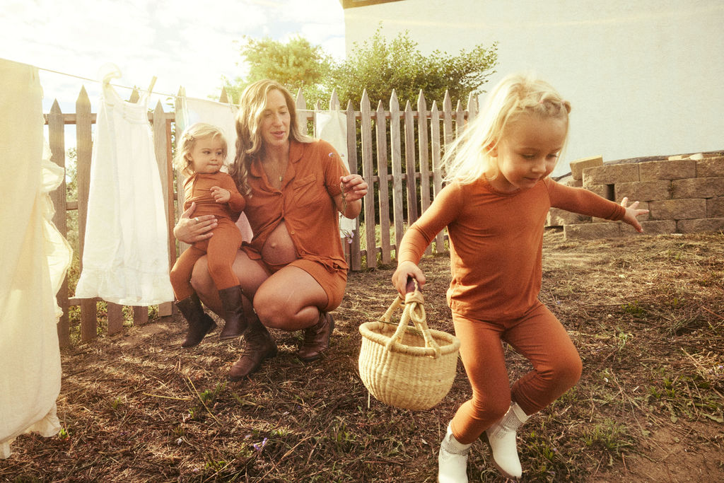 Woman and two children in matching terracotta pajamas outdoors with a wooden fence and clothesline in the background.