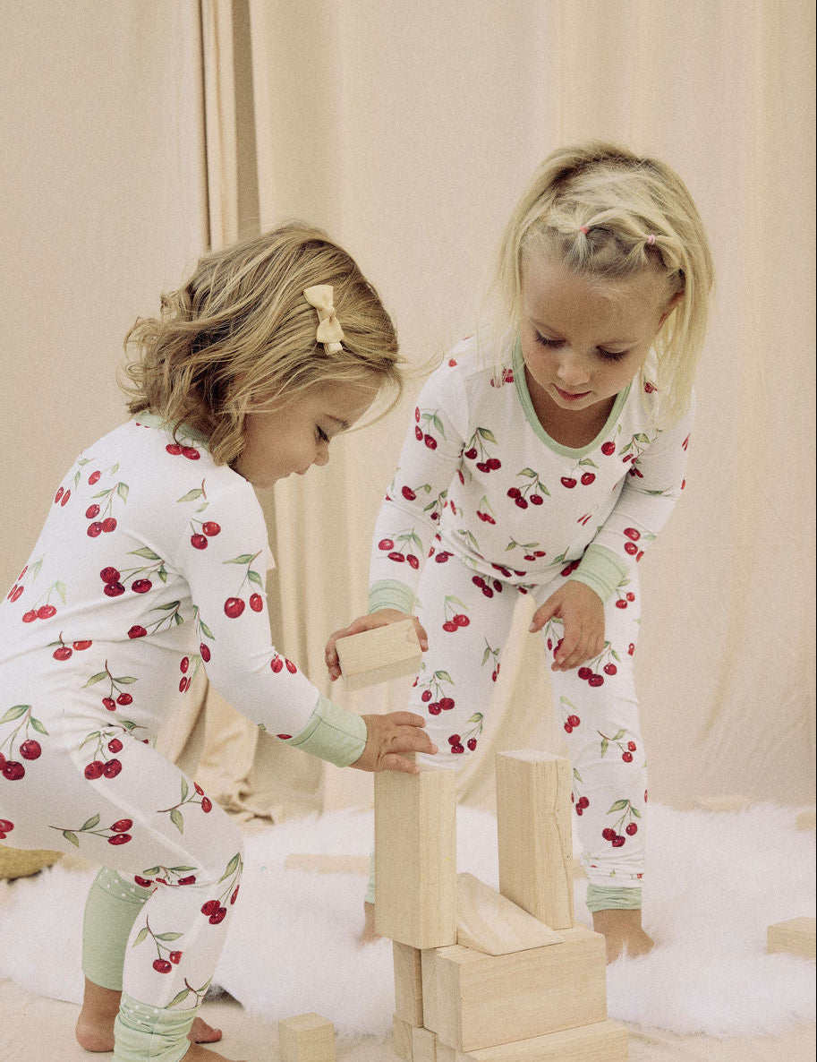 Two children in matching cherry pajamas playing with wooden blocks on a light-colored floor.