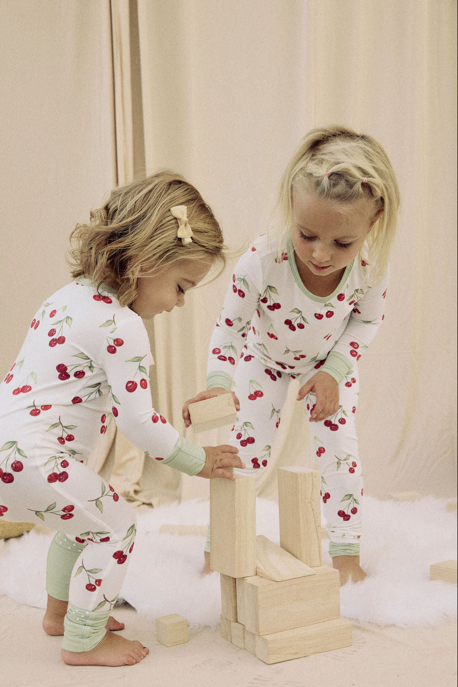Two children in matching cherry pajamas playing with wooden blocks on a light-colored floor.