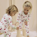 Two children in matching cherry pajamas playing with wooden blocks on a light-colored floor.