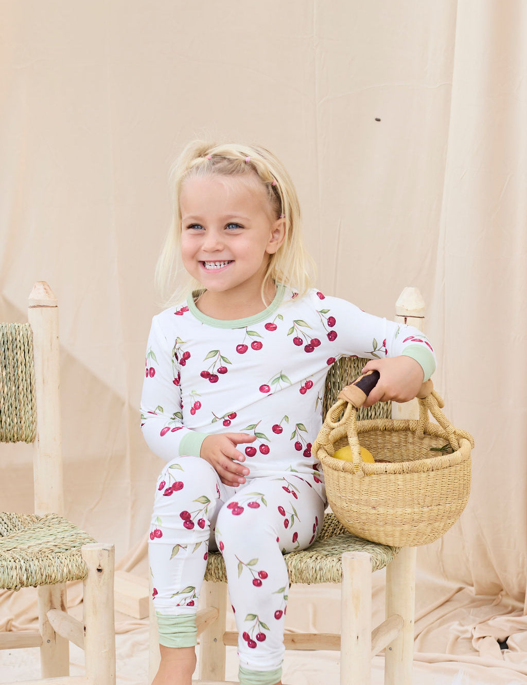 Child wearing hand-painted cherry-patterned pajamas sitting on a wooden stool holding a basket.