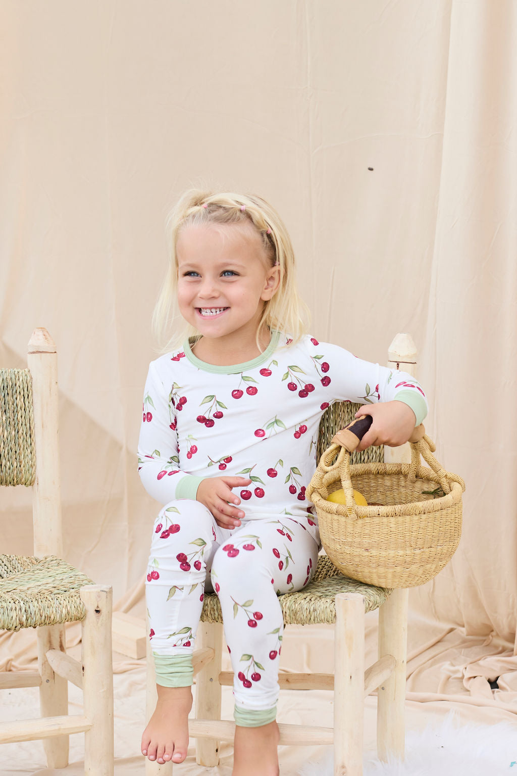 Child wearing hand-painted cherry-patterned pajamas sitting on a wooden stool holding a basket.