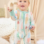 Baby in a hand-painted colorful surfboard onesie standing in a cozy room with soft lighting.