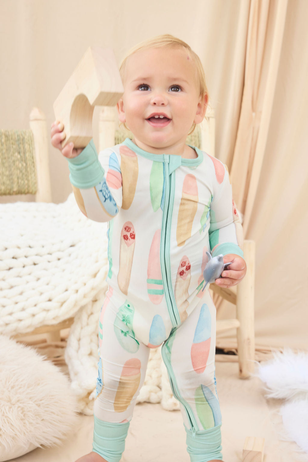 Baby in a hand-painted colorful surfboard onesie standing in a cozy room with soft lighting.
