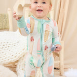 Baby in a hand-painted colorful surfboard onesie standing in a cozy room with soft lighting.