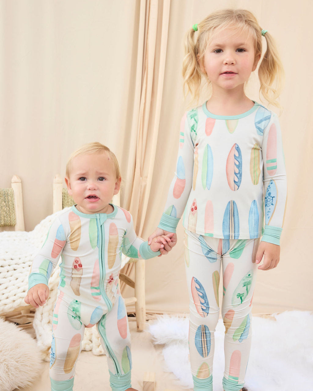 Two children wearing matching hand-painted pajamas with a colorful pastel surfboard pattern in a cozy room.