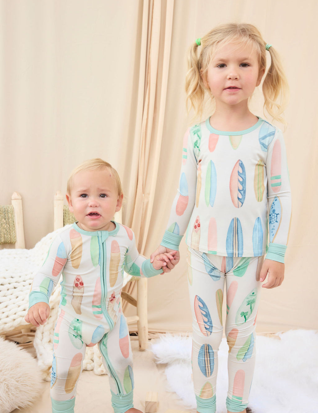 Two children wearing matching hand-painted pajamas with a colorful pastel surfboard pattern in a cozy room.