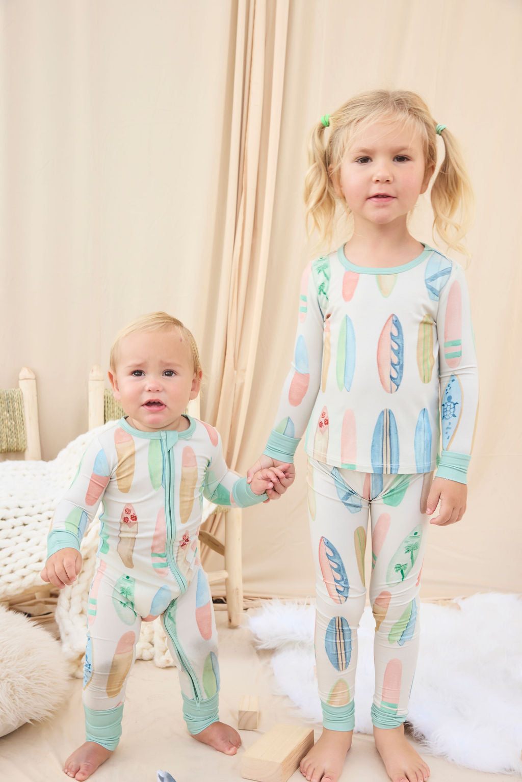Two children wearing matching hand-painted pajamas with a colorful pastel surfboard pattern in a cozy room.