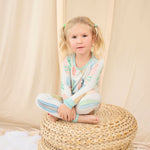 Young girl in hand-painted pastel surfboard pajamas sitting on a woven ottoman against a beige curtain.