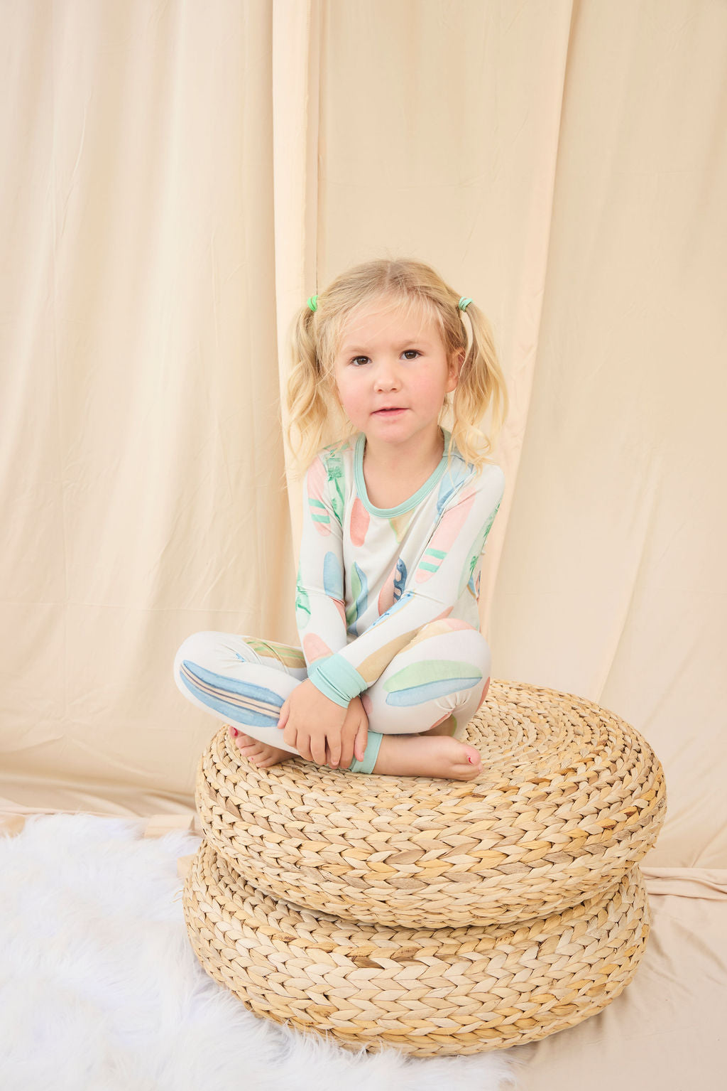 Young girl in hand-painted pastel surfboard pajamas sitting on a woven ottoman against a beige curtain.
