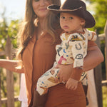 Woman holding a baby wearing a cowboy print onesie outdoors with a wooden fence and greenery in the background