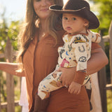 Woman holding a baby wearing a cowboy print onesie outdoors with a wooden fence and greenery in the background