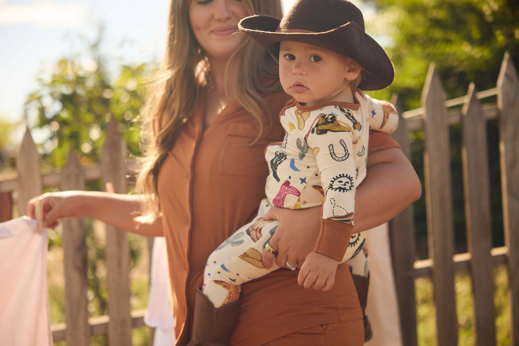 Woman holding a baby wearing a cowboy print onesie outdoors with a wooden fence and greenery in the background