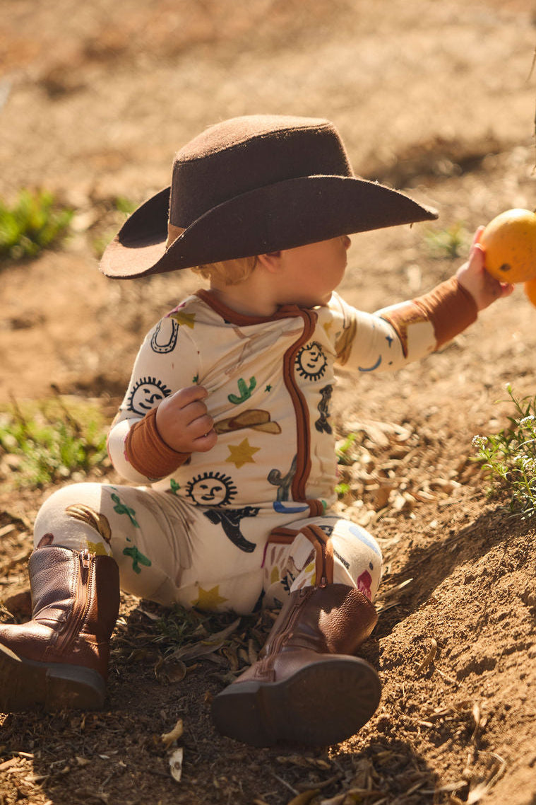 Child in cowboy pajamas onesie with a cowboy hat and boots, picking an orange, on a dirt ground.