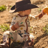 Child in cowboy pajamas onesie with a cowboy hat and boots, picking an orange, on a dirt ground.