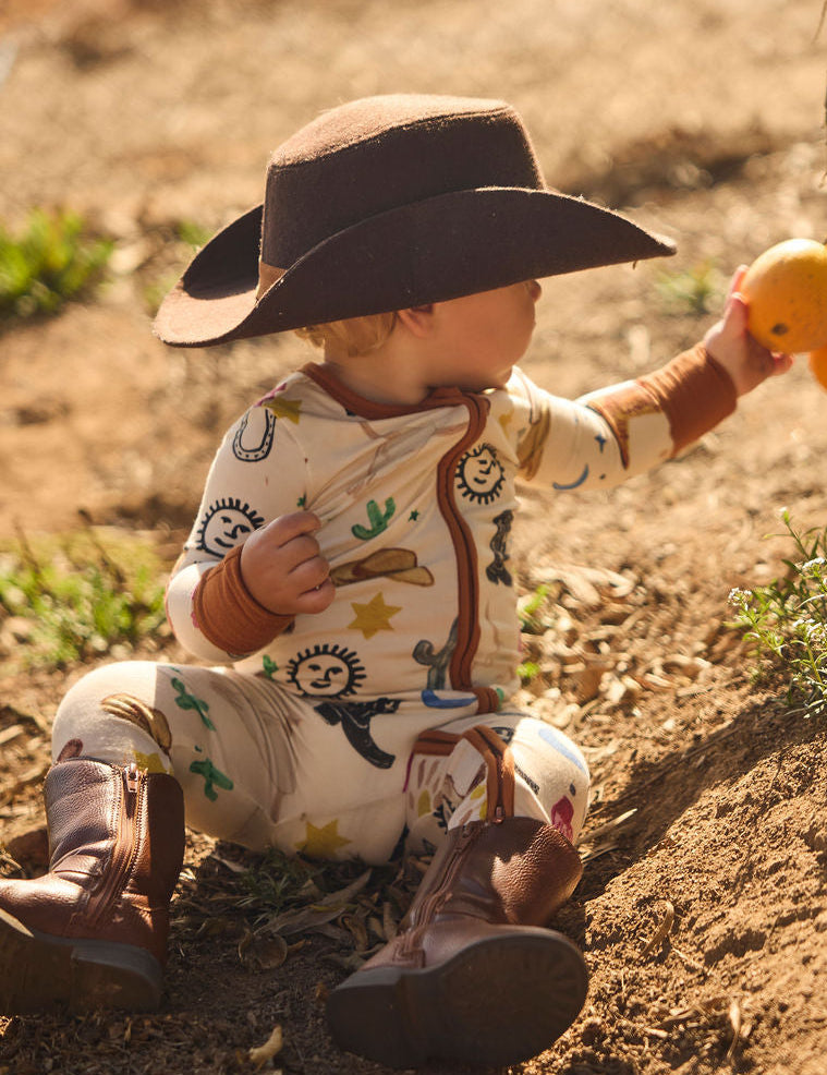 Child in cowboy pajamas onesie with a cowboy hat and boots, picking an orange, on a dirt ground.