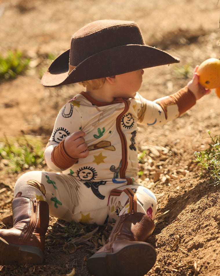 Child in cowboy pajamas onesie with a cowboy hat and boots, picking an orange, on a dirt ground.