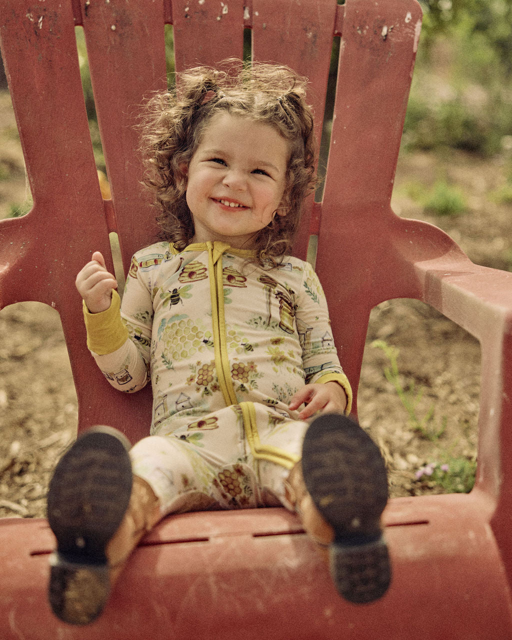 Child sitting on a red chair outdoors wearing a hand-painted honey and bees themed pajama with a blurred natural background