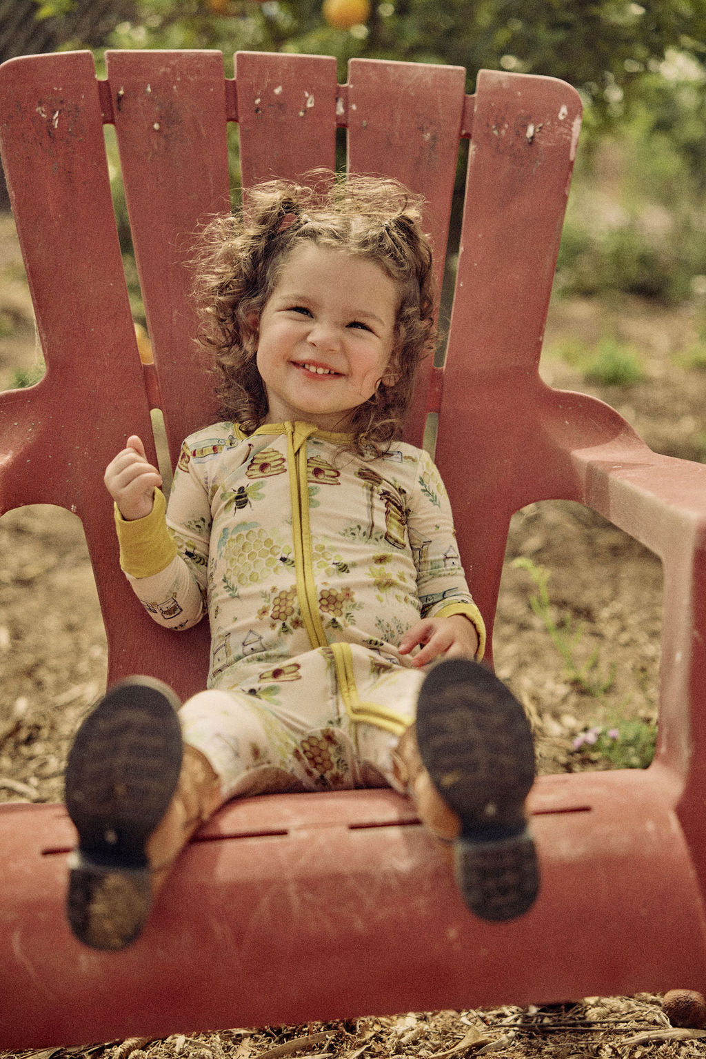 Child sitting on a red chair outdoors wearing a hand-painted honey and bees themed pajama with a blurred natural background