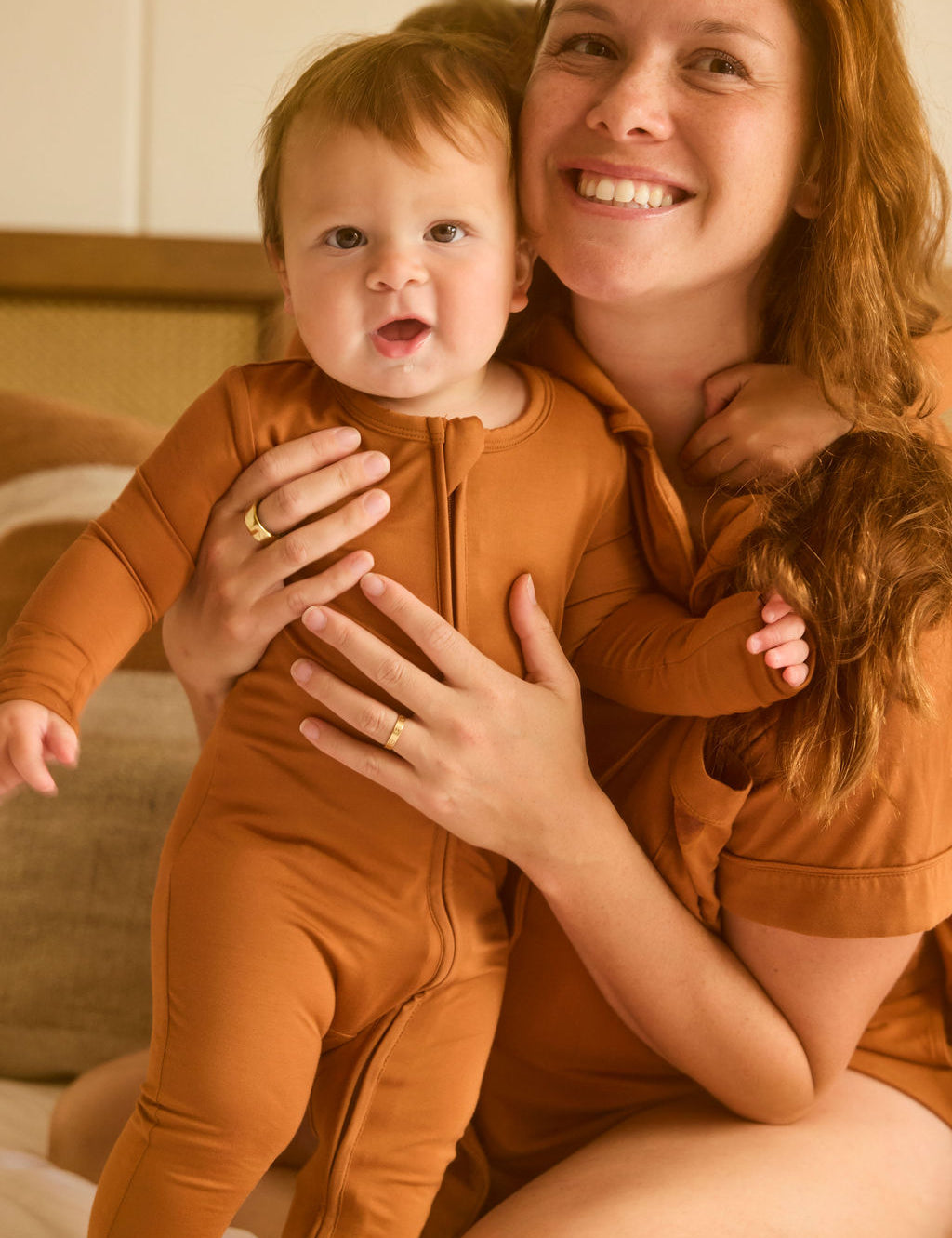 Woman holding a baby in matching terracotta pajama set indoors.