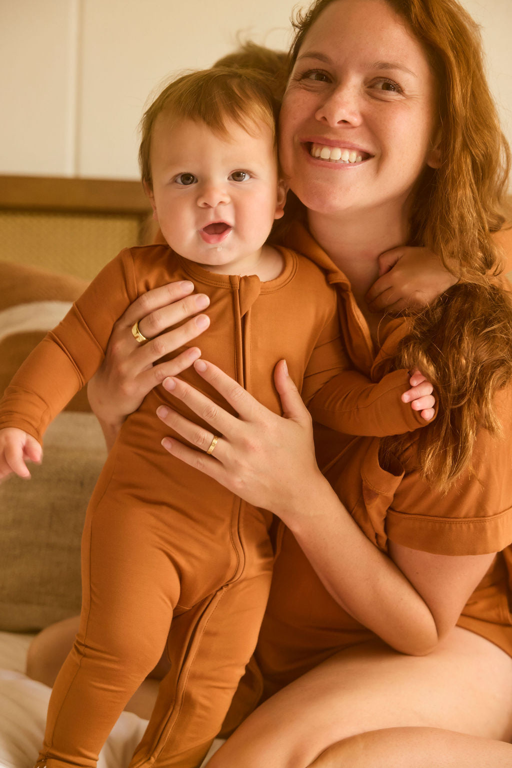 Woman holding a baby in matching terracotta pajama set indoors.