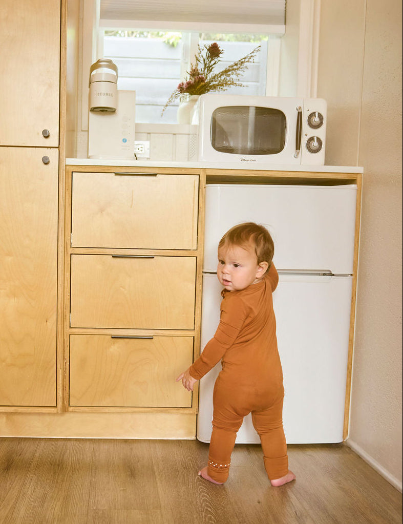Child in a terracotta onesie standing in a kitchen with wooden cabinets and a white refrigerator.