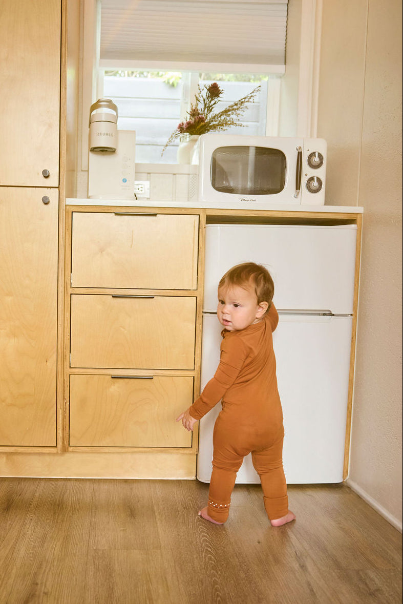 Child in a terracotta onesie standing in a kitchen with wooden cabinets and a white refrigerator.
