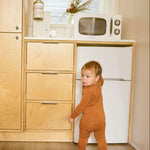 Child in a terracotta onesie standing in a kitchen with wooden cabinets and a white refrigerator.