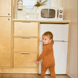 Child in a terracotta onesie standing in a kitchen with wooden cabinets and a white refrigerator.