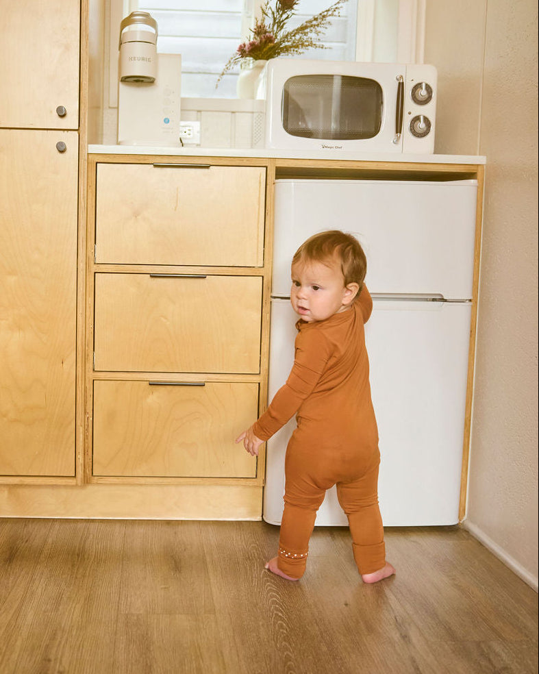 Child in a Canyon Clay Onesie standing in a kitchen with wooden cabinets and a white refrigerator.