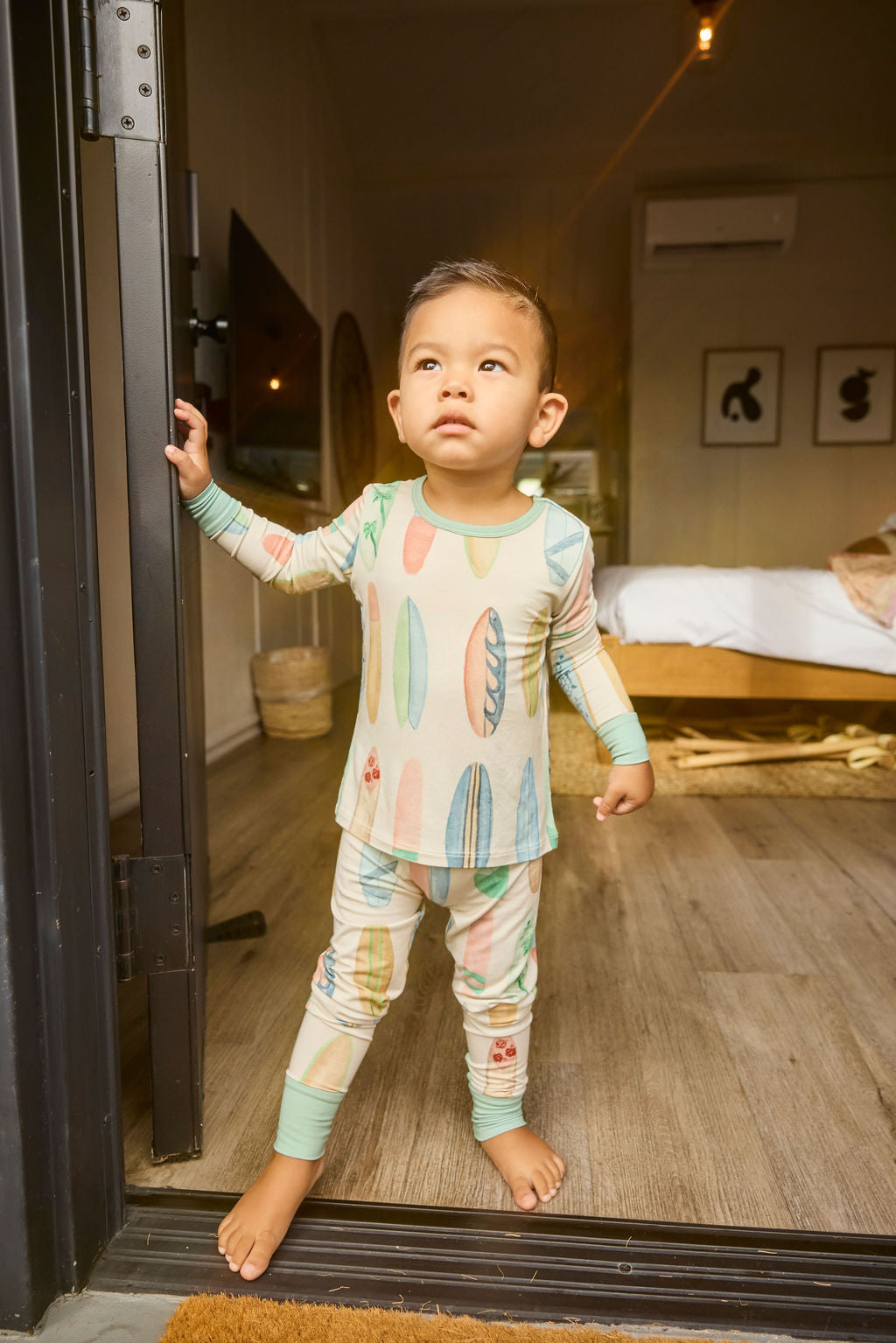 Child in hand-painted surfboard pajamas standing in a doorway with a bedroom in the background