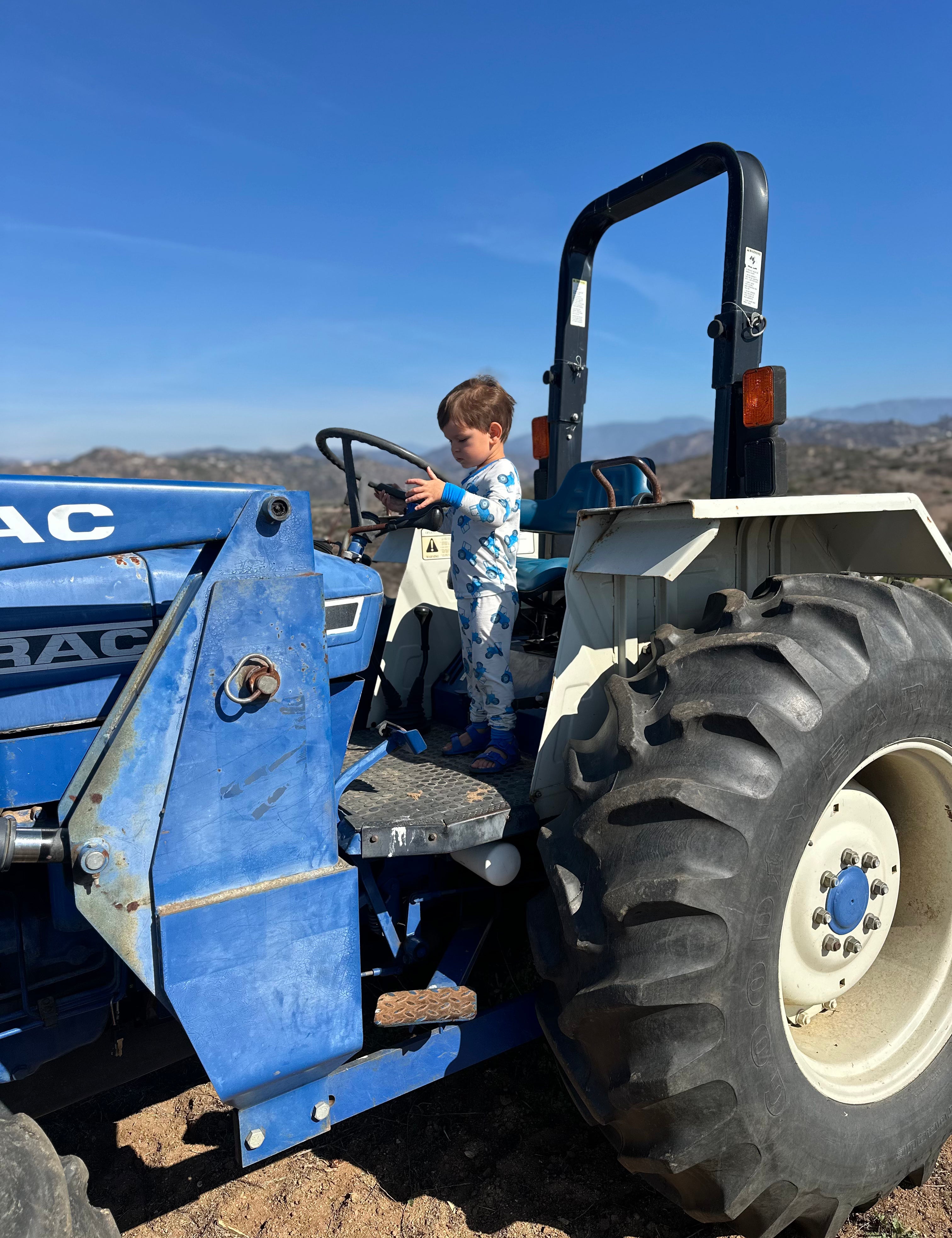 Child standing on a blue tractor  with a blue tractor pajama set with a clear blue sky in the background