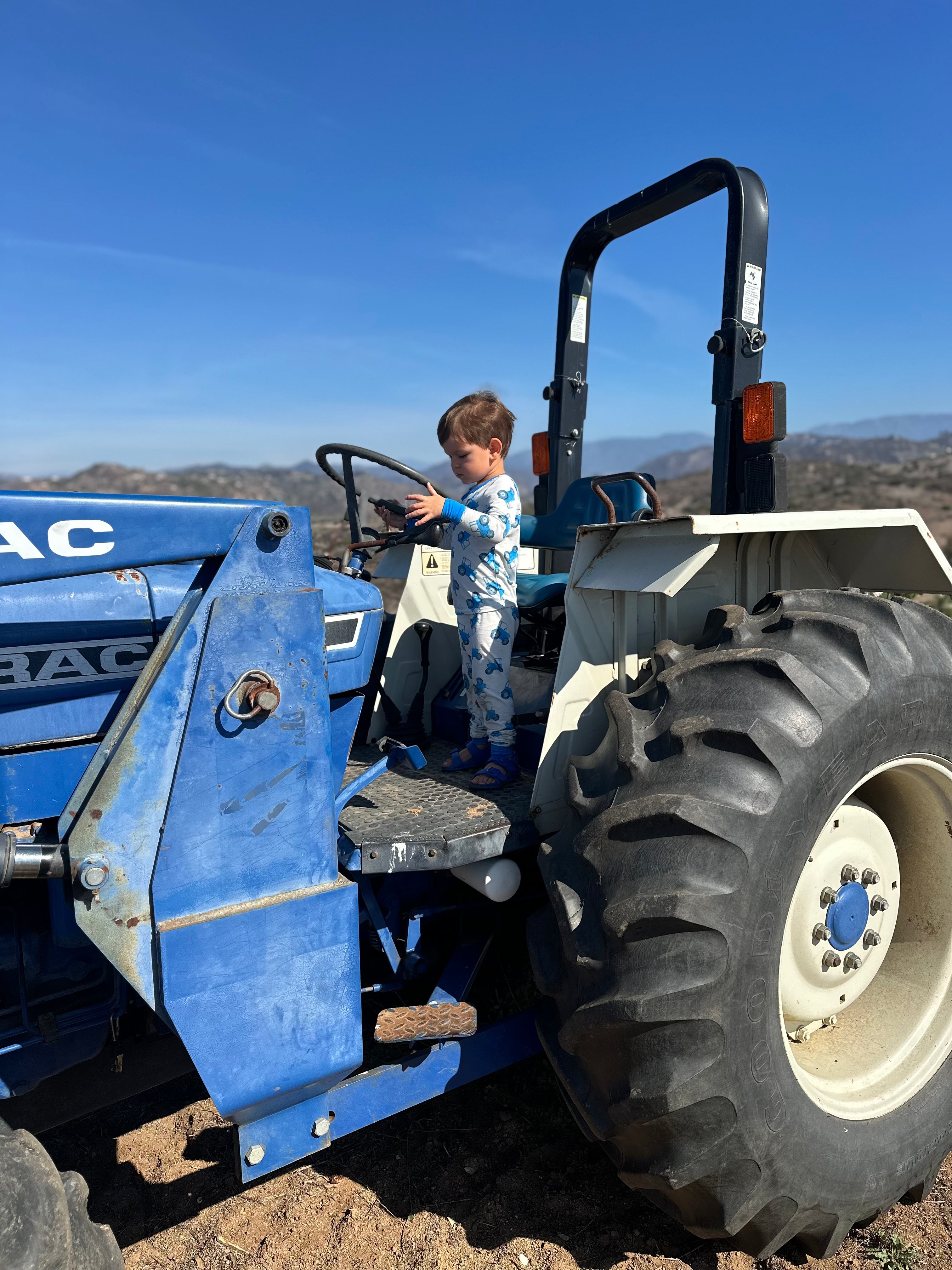 Child standing on a blue tractor  with a blue tractor pajama set with a clear blue sky in the background