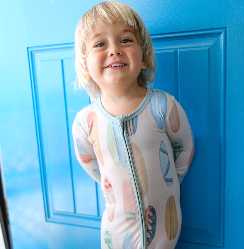 Child wearing a hand-painted surfboard onesie standing in front of a blue door.