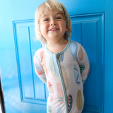 Child wearing a hand-painted surfboard onesie standing in front of a blue door.