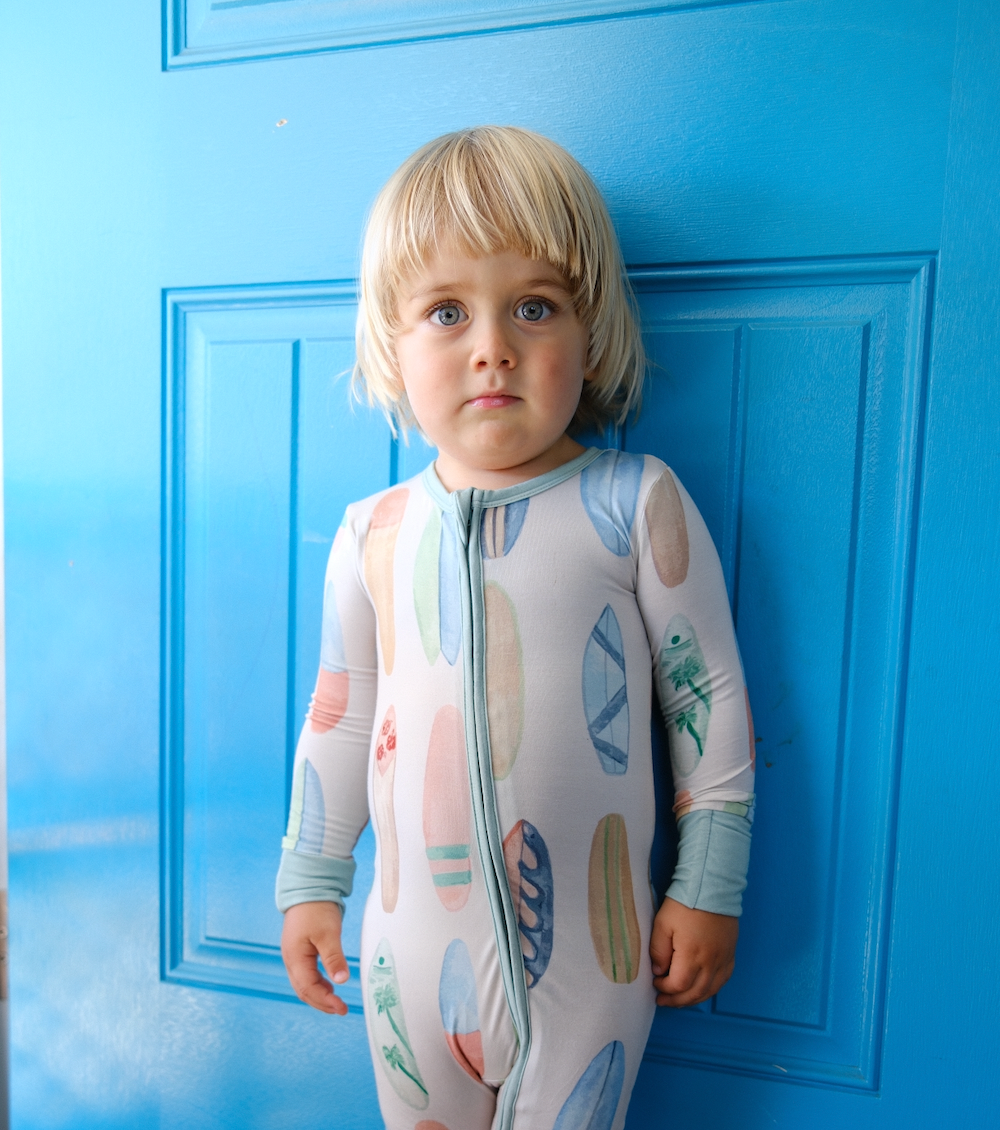 Child wearing a hand-painted surboard onesie standing against a blue door
