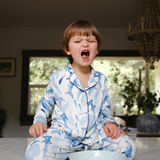 Child in blue pajamas sitting on a floor with a bowl, in a room with a chandelier and window.