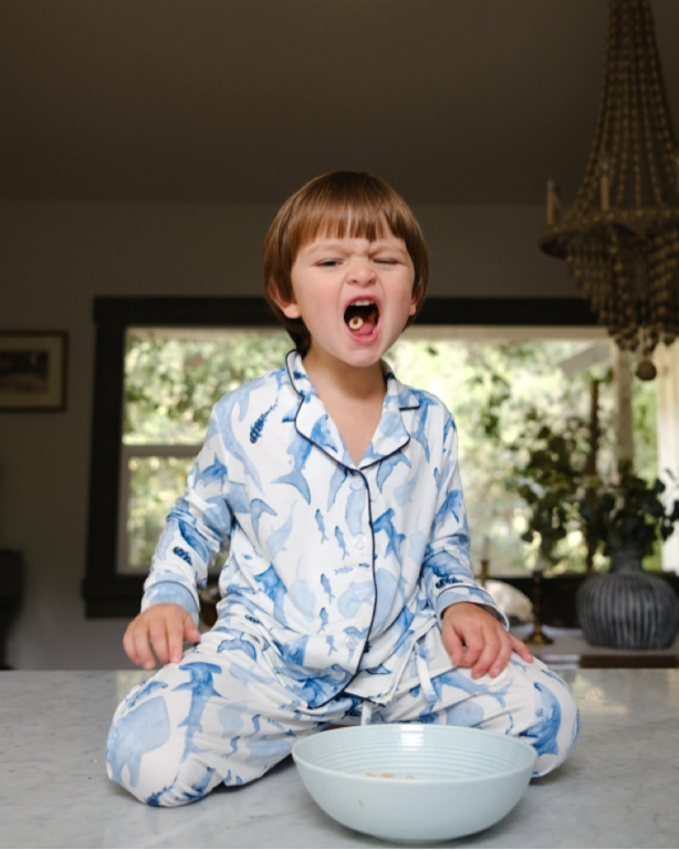 Child in blue pajamas sitting on a floor with a bowl, in a room with a chandelier and window.