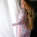 Woman wearing a pink pj set with colorful patterns standing in front of a white curtain.